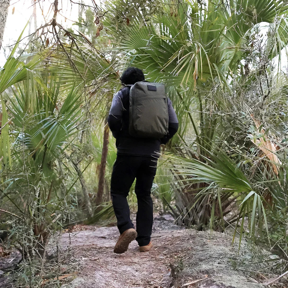 Person rucking with a GORUCK backpack on a forest trail, surrounded by green palm trees.