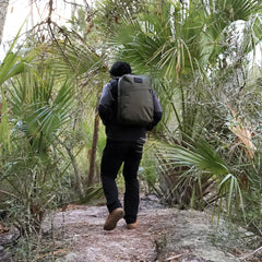 Person rucking with a GORUCK backpack on a forest trail, surrounded by green palm trees.