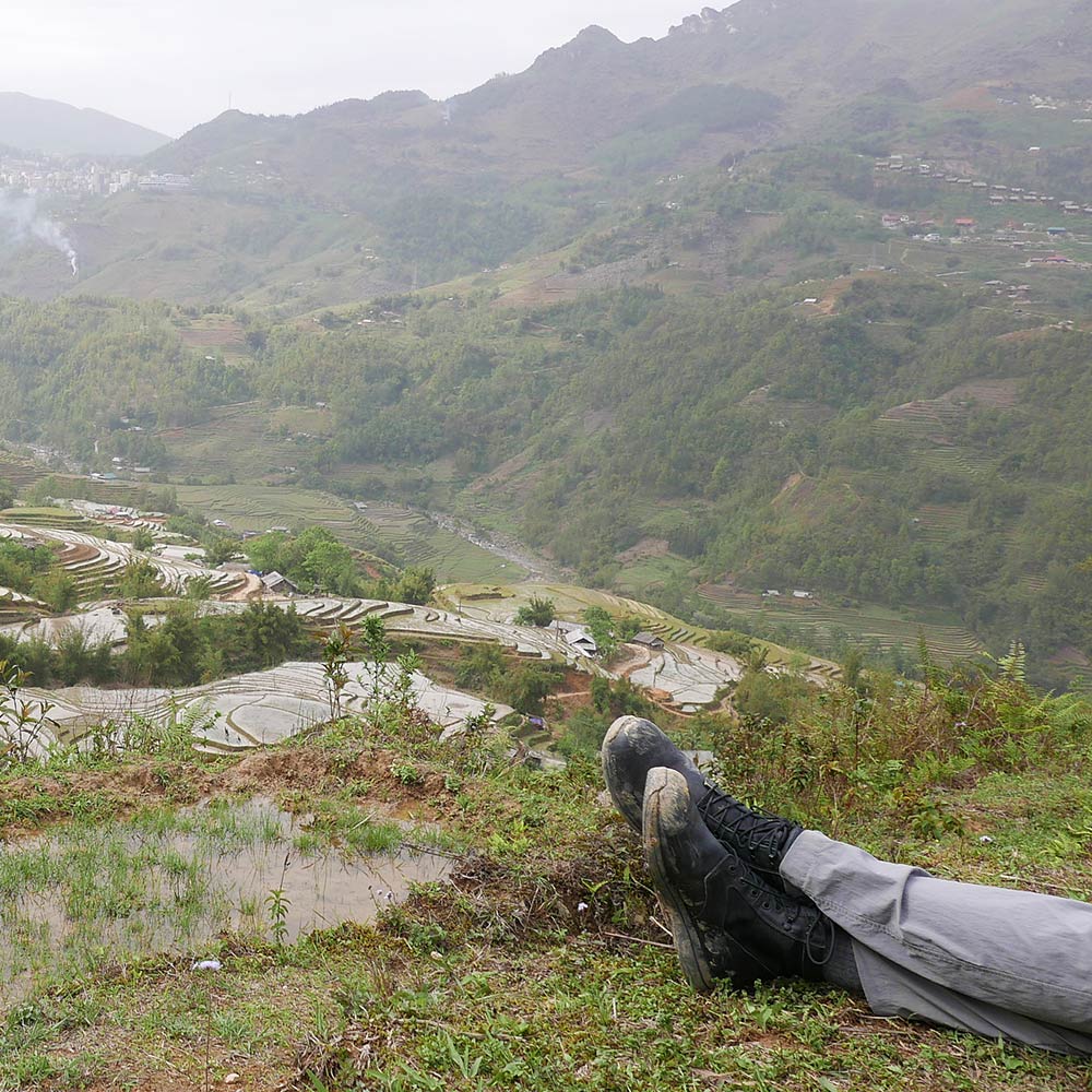 Person lying on grass with boots visible, overlooking terraced fields and hills under a cloudy sky.