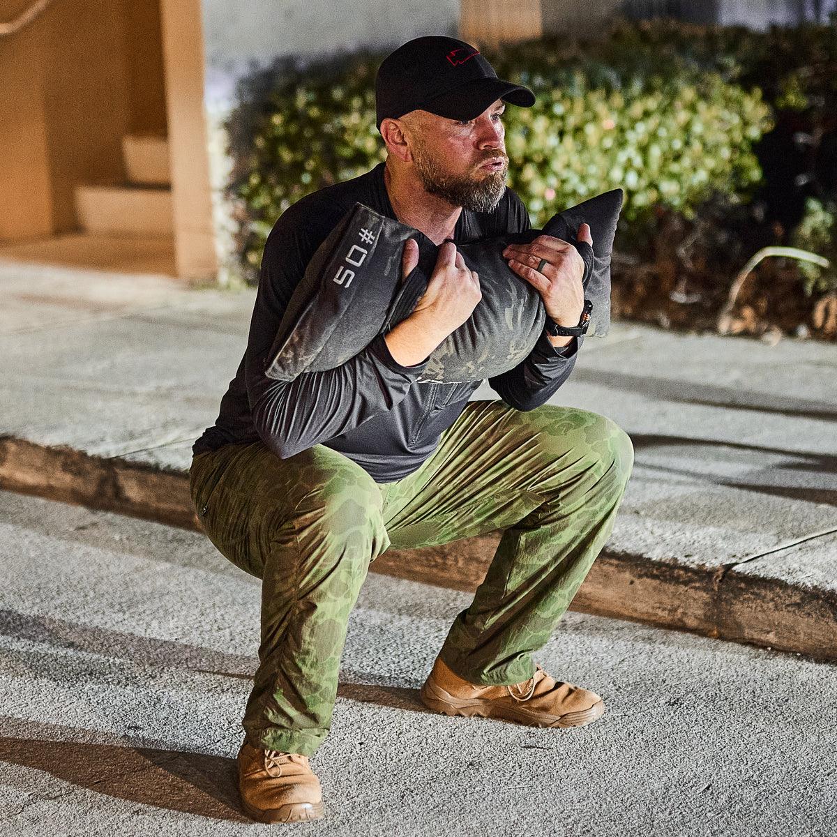 Man wearing GORUCK performance half zip and camo pants squatting with a 50 lb sandbag outdoors