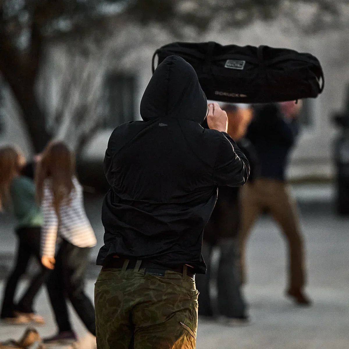Man in GORUCK sun hoodie tossing a black ruck bag outdoors during group rucking activity
