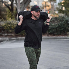 Man outdoors wearing GORUCK black Sun Hoodie, camo pants, carrying weighted ruck sack