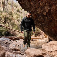 Man hiking outdoors in rugged gear, wearing olive green pants, black jacket, and backpack