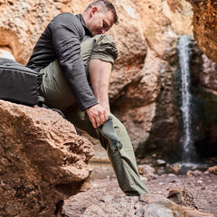 Man outdoors near waterfall putting on green Merino wool socks, GORUCK rucking gear shown