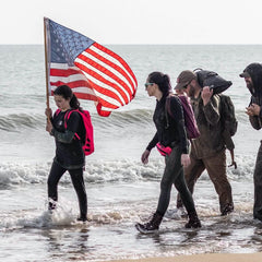 GORUCK team rucking on the beach, American flag, backpacks, ocean waves, outdoor gear