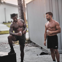 Two muscular men in GORUCK shorts holding drinks outdoors after a workout, smiling and relaxed.