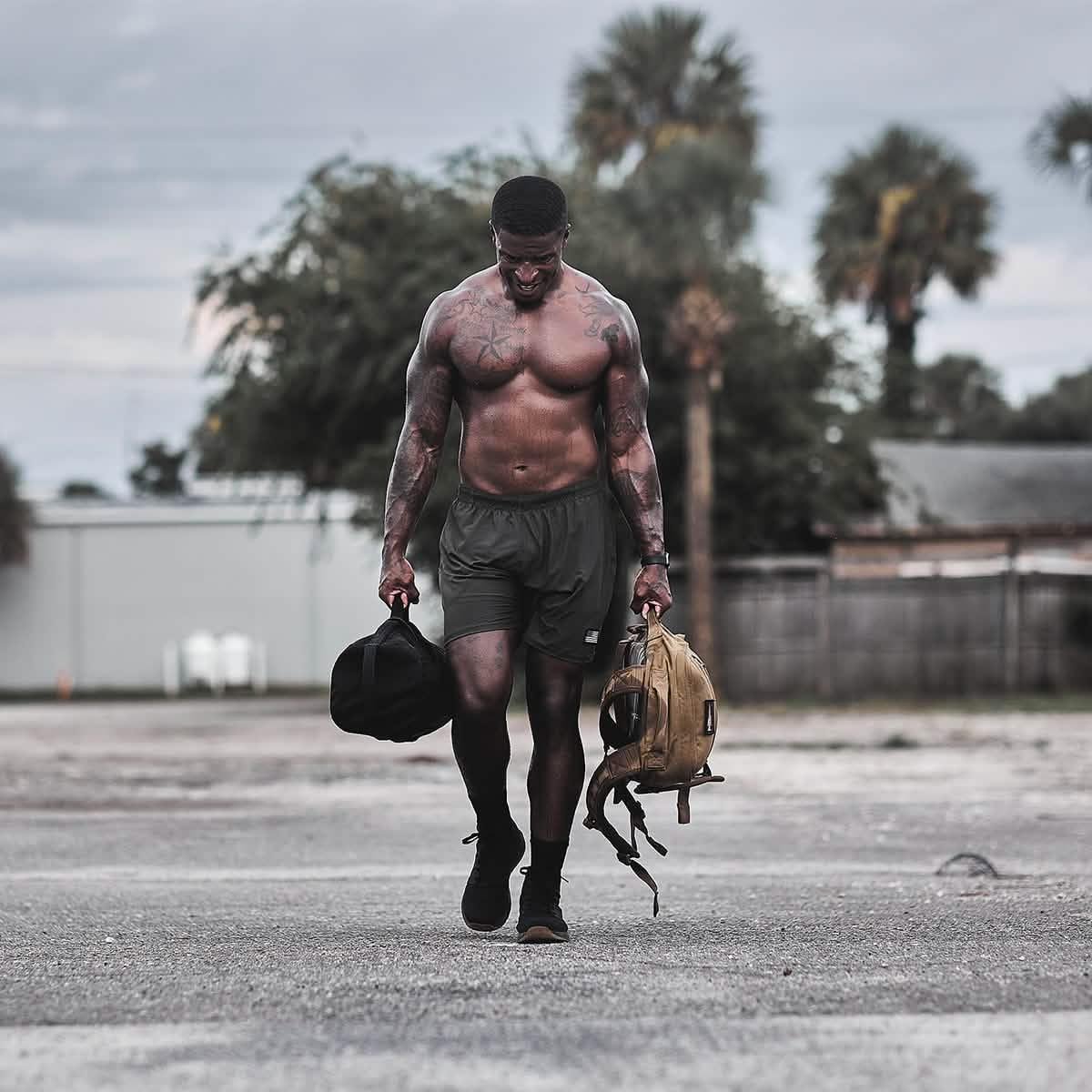 Athletic man rucking outdoors with GORUCK sandbag and backpack, palm trees in background.