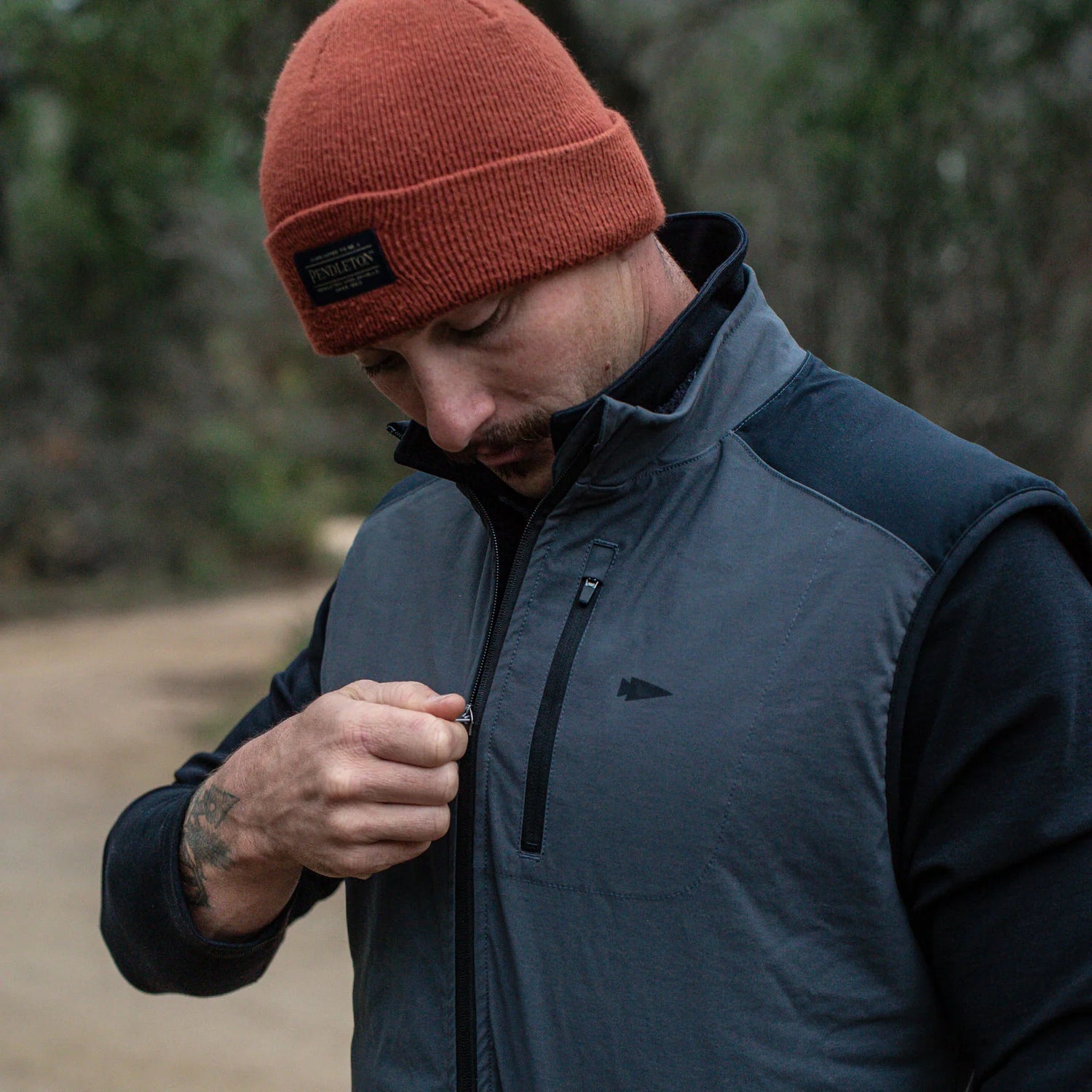 Man outdoors zipping GORUCK jacket, wearing rust beanie, durable rucking apparel