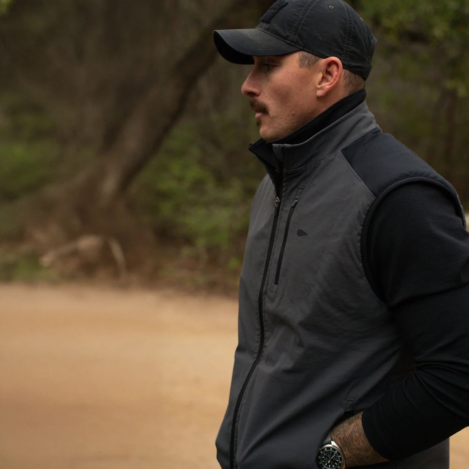 Man wearing GORUCK tactical vest and cap outdoors on a dirt path, forest background