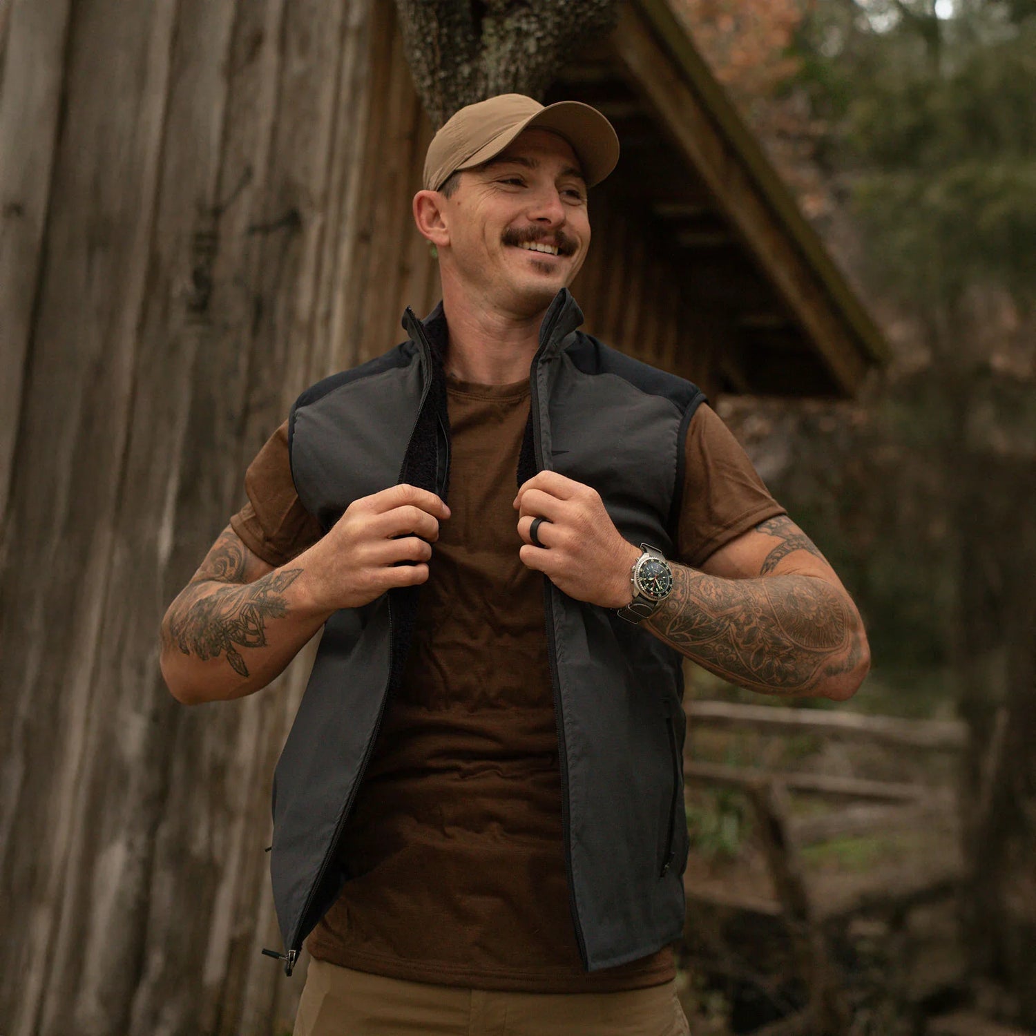 Man wearing GORUCK tactical vest and cap outdoors, rustic cabin background, rucking gear