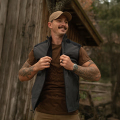 Man wearing GORUCK tactical vest and cap outdoors, rustic cabin background, rucking gear