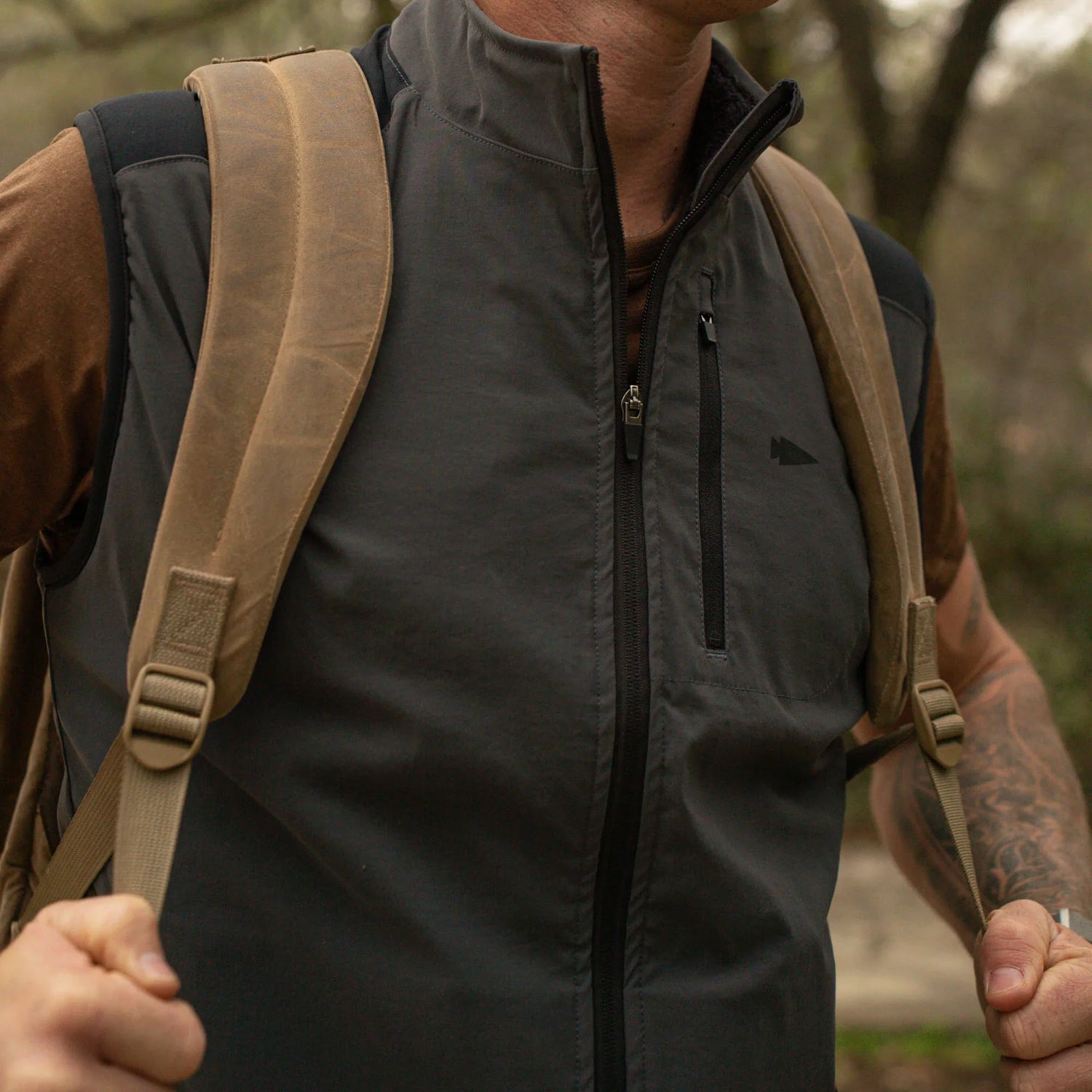 Man wearing GORUCK rucking vest and tan backpack outdoors, gripping shoulder straps