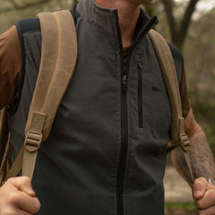Man wearing GORUCK rucking vest and tan backpack outdoors, gripping shoulder straps