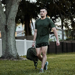 Man wearing olive GORUCK Tough Mesh Tee, carrying rucksack outdoors on grass