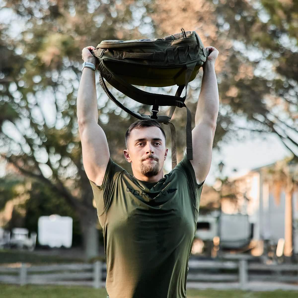 Man outdoors lifting rucksack overhead wearing olive GORUCK Tough Mesh Tee