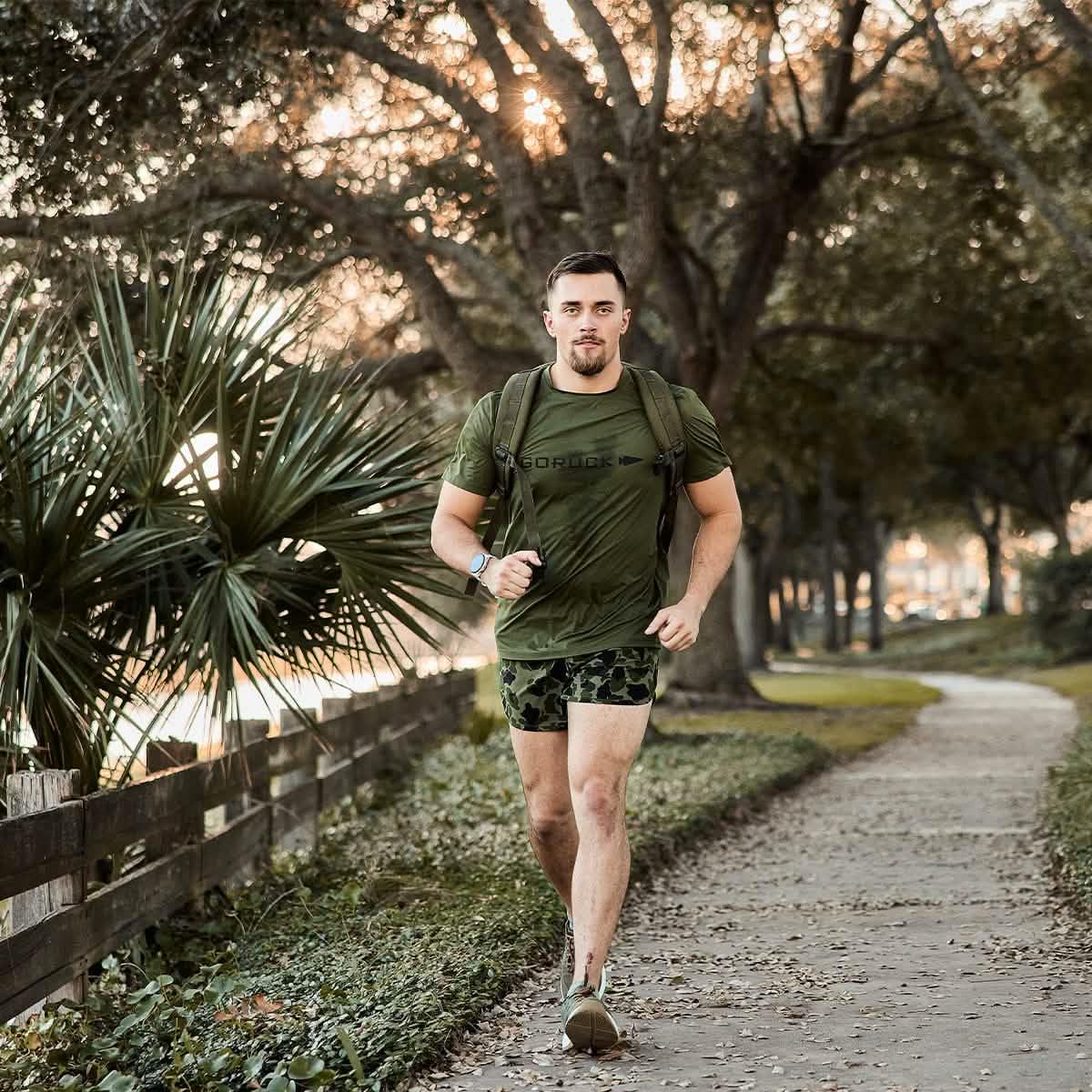 Man rucking in GORUCK olive Tough Mesh Tee outdoors, wearing rucksack on tree-lined path