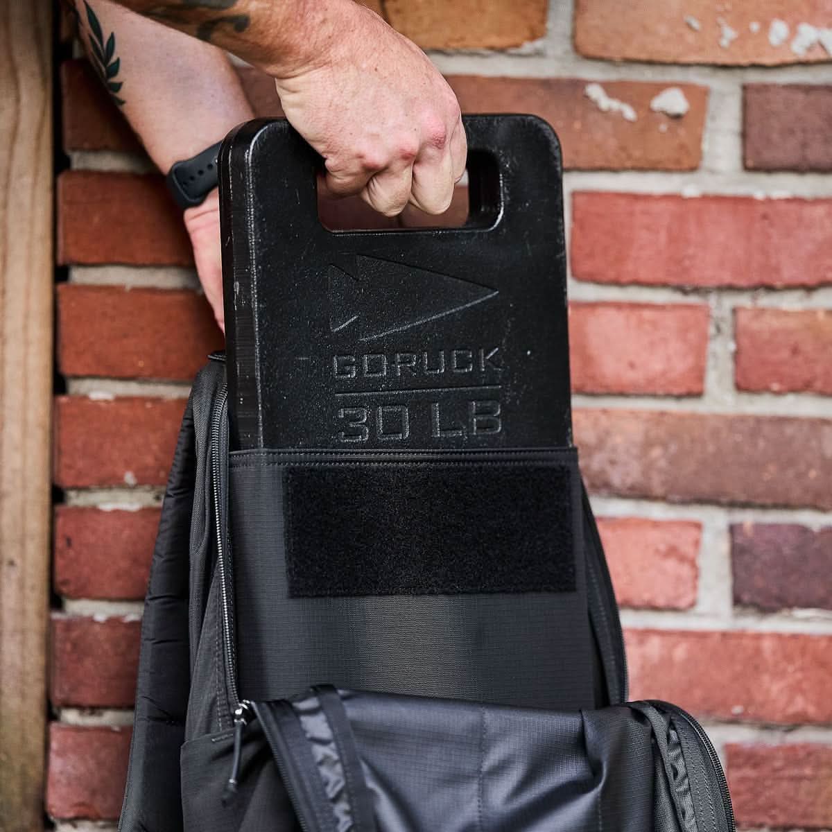 A hand places a black ruck plate into the Basic Rucker backpack in front of a brick wall, ready for your next rucking adventure.