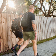 Two men rucking outdoors with GORUCK backpacks, walking on a sidewalk by a wooden fence