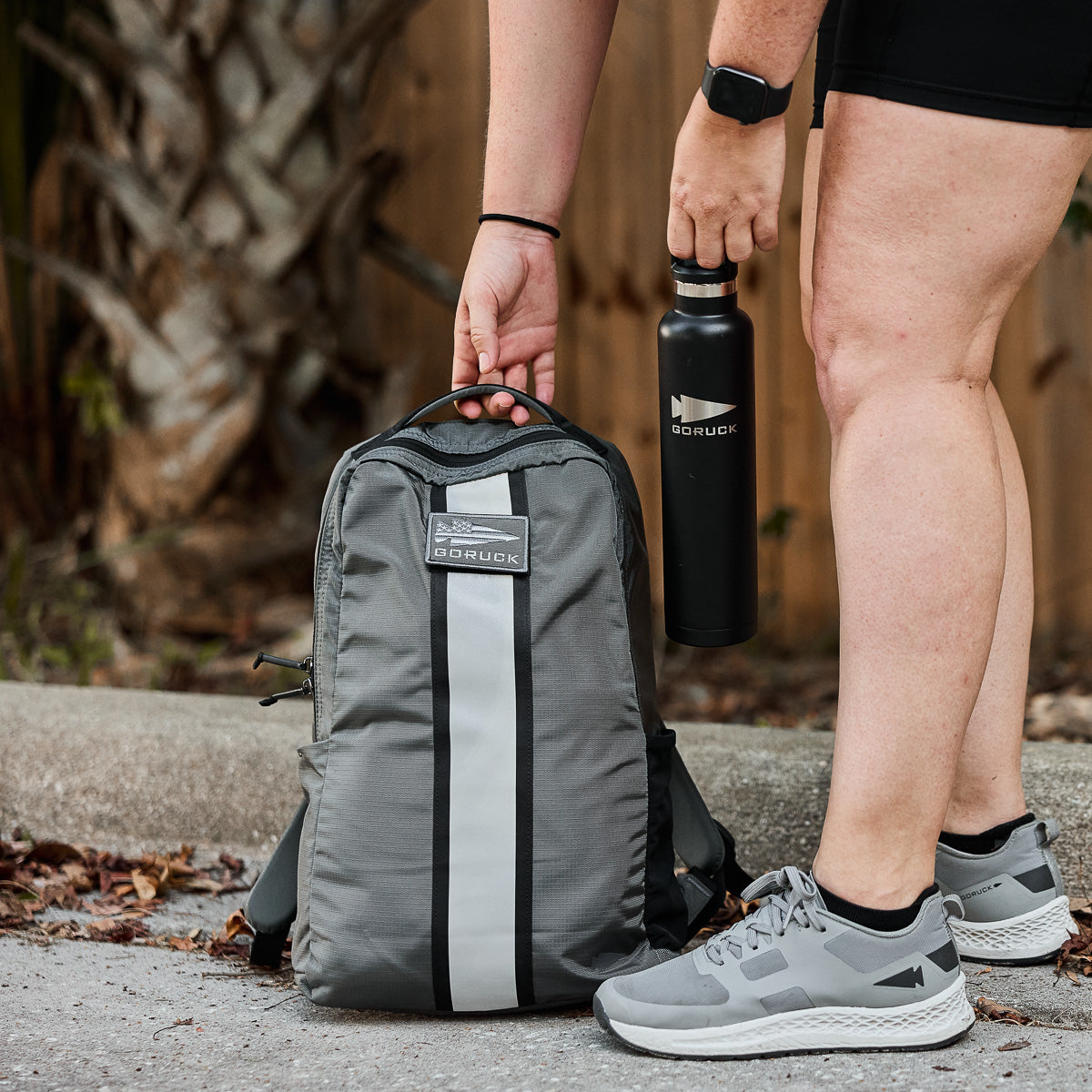 Person in shorts stands by a gray backpack and holds a black water bottle outdoors on a sidewalk.