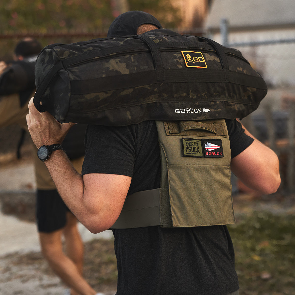 A person carries a heavy sandbag outdoors while wearing the Training Weight Vest 2.0, featuring patches on the back.
