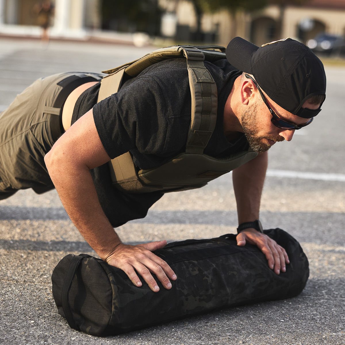 A man wears the Training Weight Vest 2.0 while doing push-ups on an exercise sandbag outdoors in sunlight.