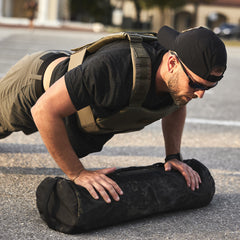 A man wears the Training Weight Vest 2.0 while doing push-ups on an exercise sandbag outdoors in sunlight.