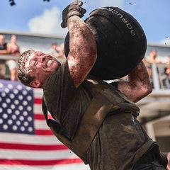 Athlete lifting GORUCK sandbag in outdoor rucking event, American flag in background