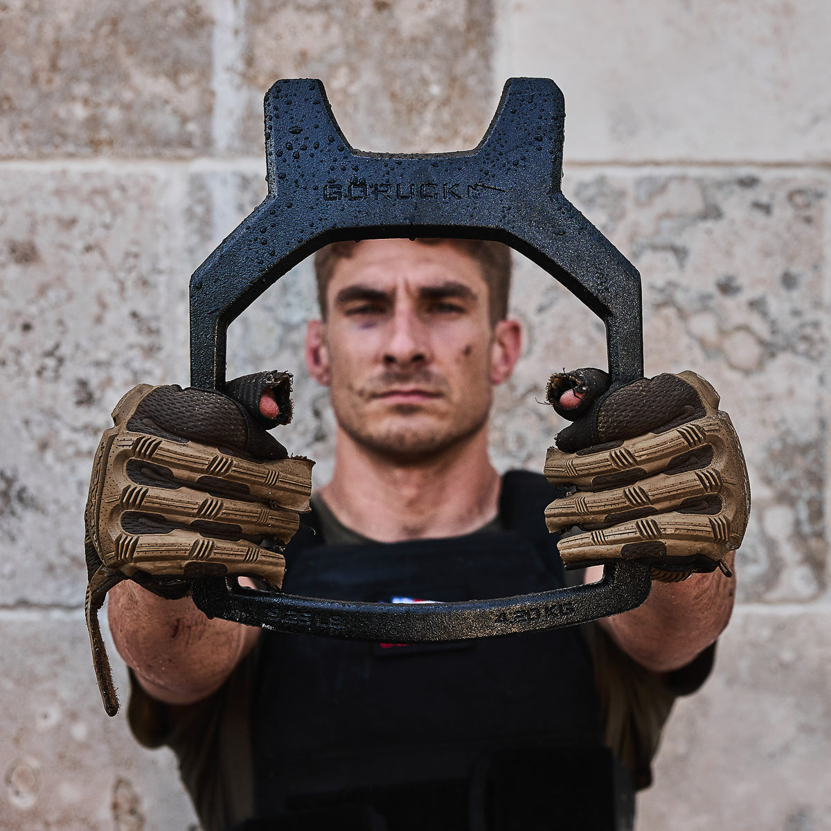 Man in tactical gloves holding a metal workout tool towards the camera, stone wall in background.