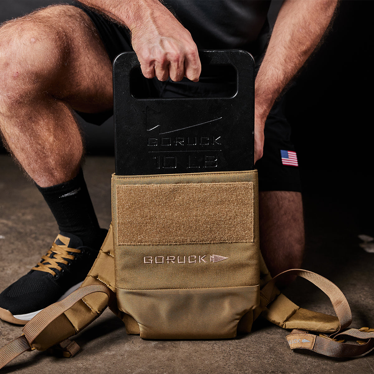 A person places a 10 lb GORUCK plate into a tan tactical vest on a concrete floor.