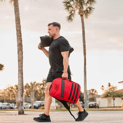 Man rucking outdoors with a red GORUCK backpack and training sandbag, palm trees in background