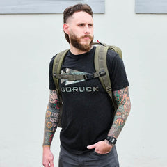 Man wearing GORUCK t-shirt and ranger green rucksack, standing outdoors.
