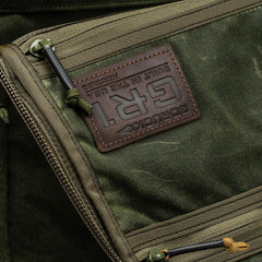 Close-up of the GR1 USA - Heritage Waxed Canvas backpack in green with a brown leather "GORUCK GR1" patch and visible zippers, highlighting classic GR1 rucksack features.