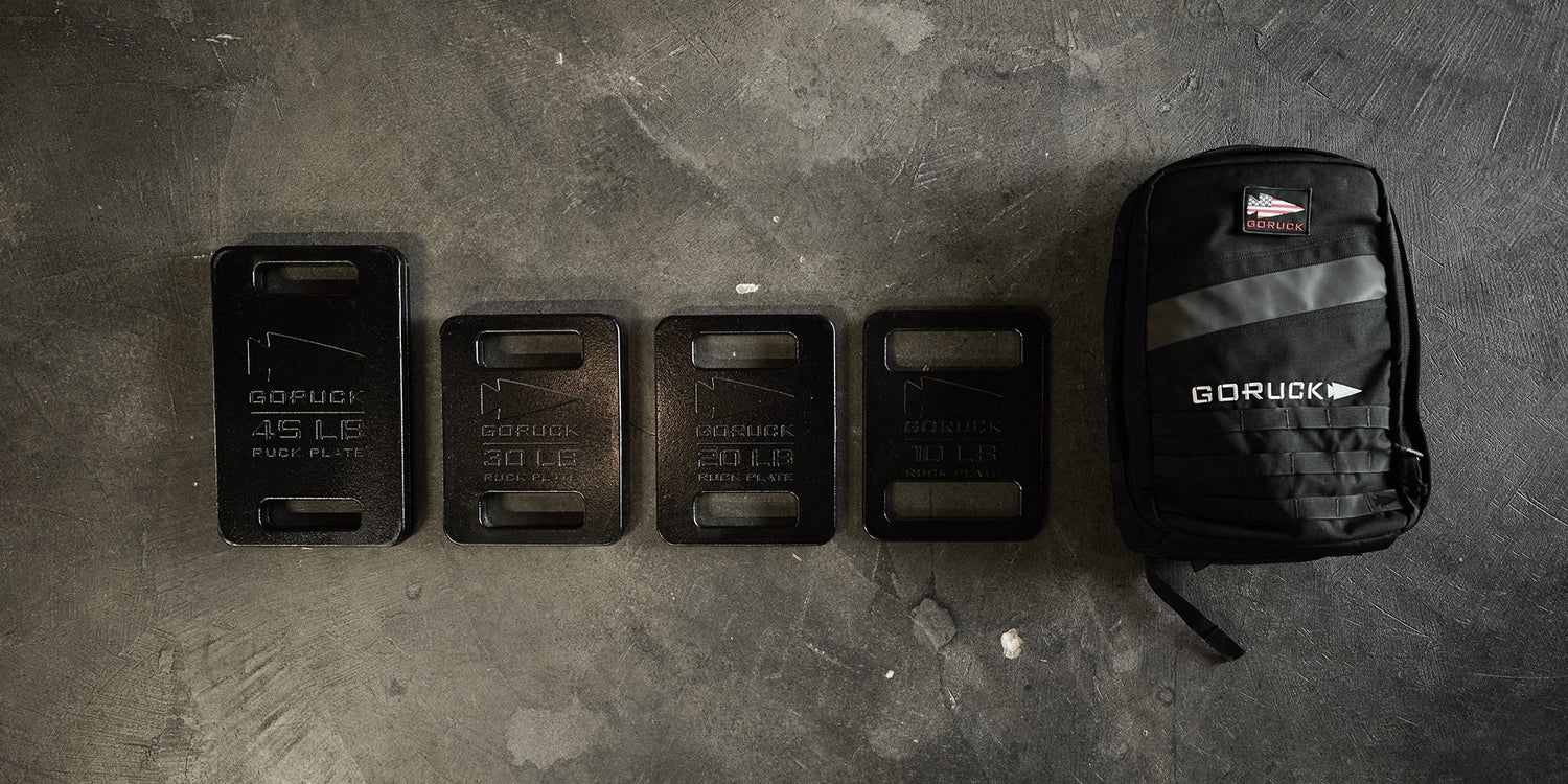 Four GORUCK weight plates and a black GORUCK backpack are lined up on a concrete floor.