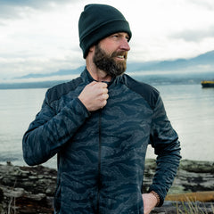 Man in GORUCK camo jacket and beanie outdoors by water with mountains in background