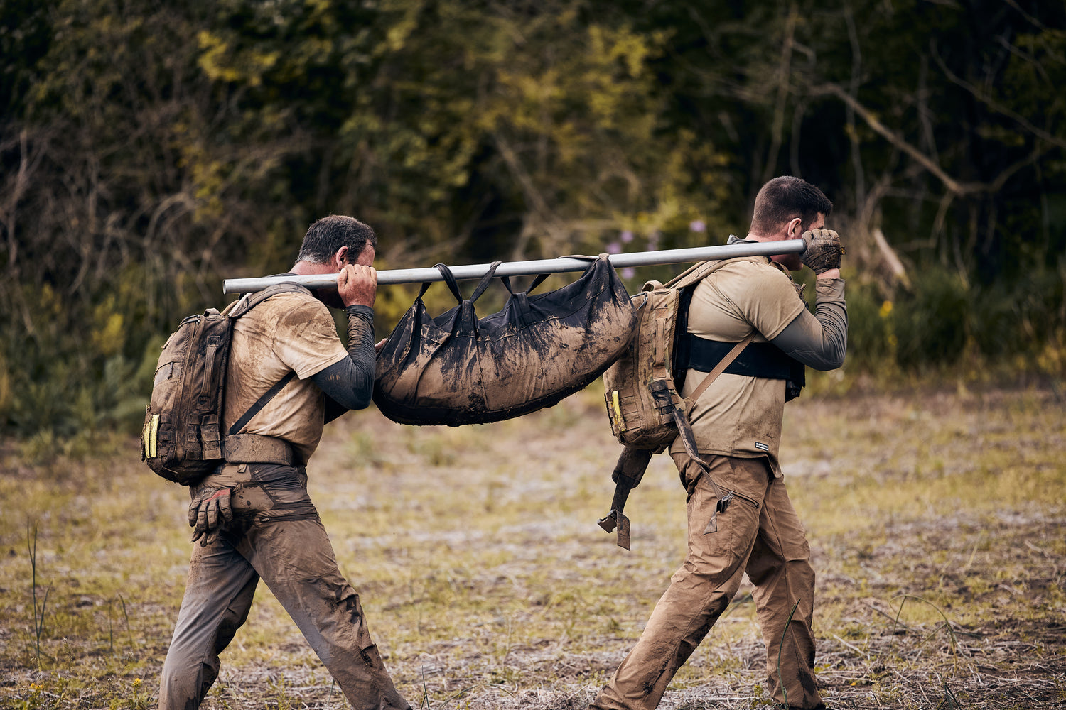 Two men carrying a muddy stretcher with gear through a field; trees and grass in the background.
