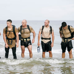 Four men in wet athletic gear, each wearing a numbered shirt and a Padded Hip Belt backpack, stand in shallow ocean water, ready for serious rucking.