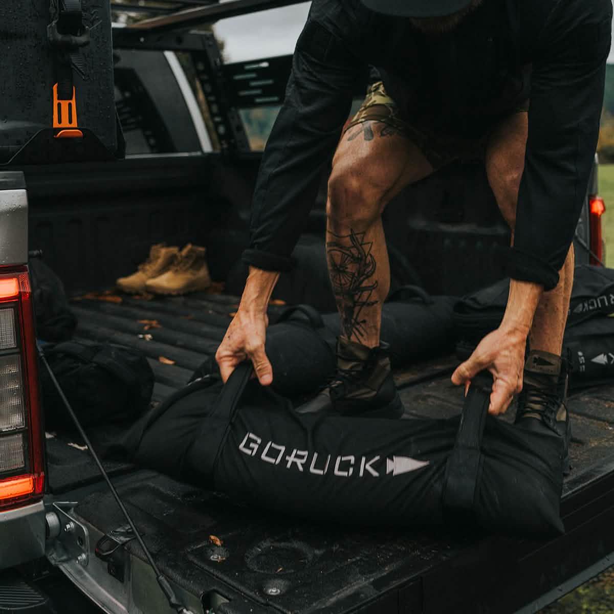 Person lifting GORUCK training sandbag from truck bed, outdoor fitness gear scene