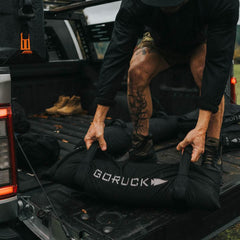 Person lifting GORUCK training sandbag from truck bed, outdoor fitness gear scene
