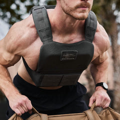 A muscular man wears the Training Weight Vest 2.0 while exercising outdoors, gripping a sandbag with both hands.
