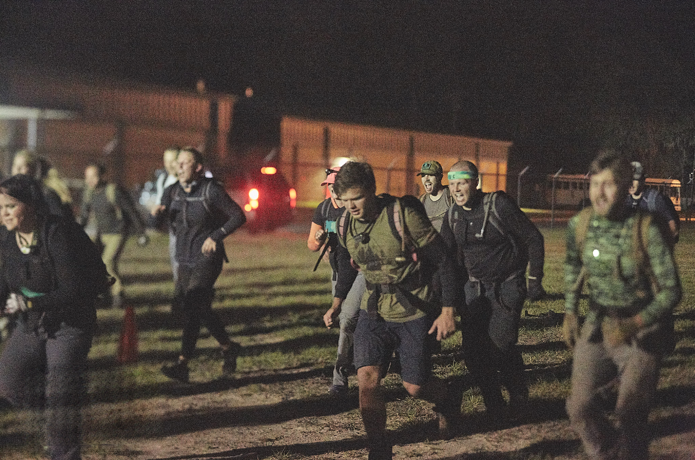 People wearing backpacks and athletic gear running outdoors at night, illuminated by artificial lights.