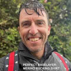 Smiling man with wet hair and red-strap backpack outdoors. Text: "Men's Rucking Shirt - Merino Wool. The perfect baselayer, made in USA.