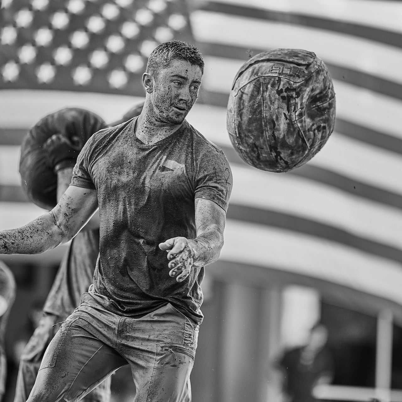 A man covered in mud throws a Sand Medicine Ball with a large American flag in the background, demonstrating core stability and strength.