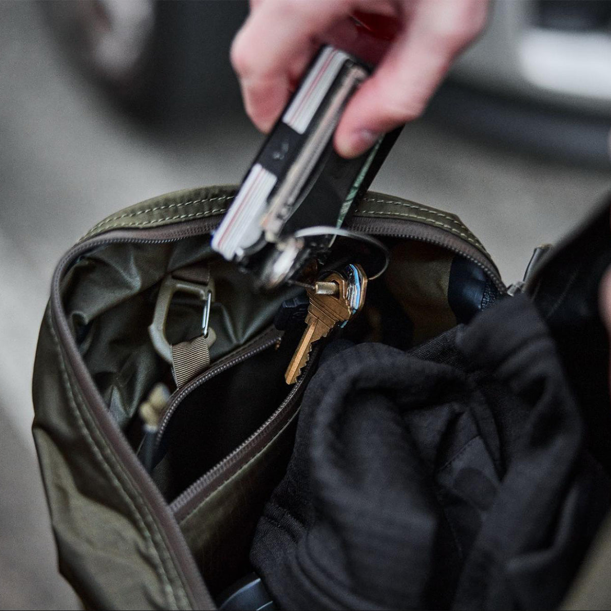 A hand places a multitool and keys into an open olive-green bag with black clothing inside.