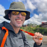 A man in a hat and sunglasses, smiling outdoors with a backpack and T-REX Hand Straps (pair)—ideal rucking gear for hand swelling relief—stands against a blurred nature background.
