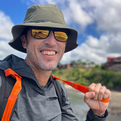 A man in a hat and sunglasses, smiling outdoors with a backpack and T-REX Hand Straps (pair)—ideal rucking gear for hand swelling relief—stands against a blurred nature background.