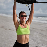 Woman in neon green GORUCK sports bra lifting rucksack overhead on beach during workout