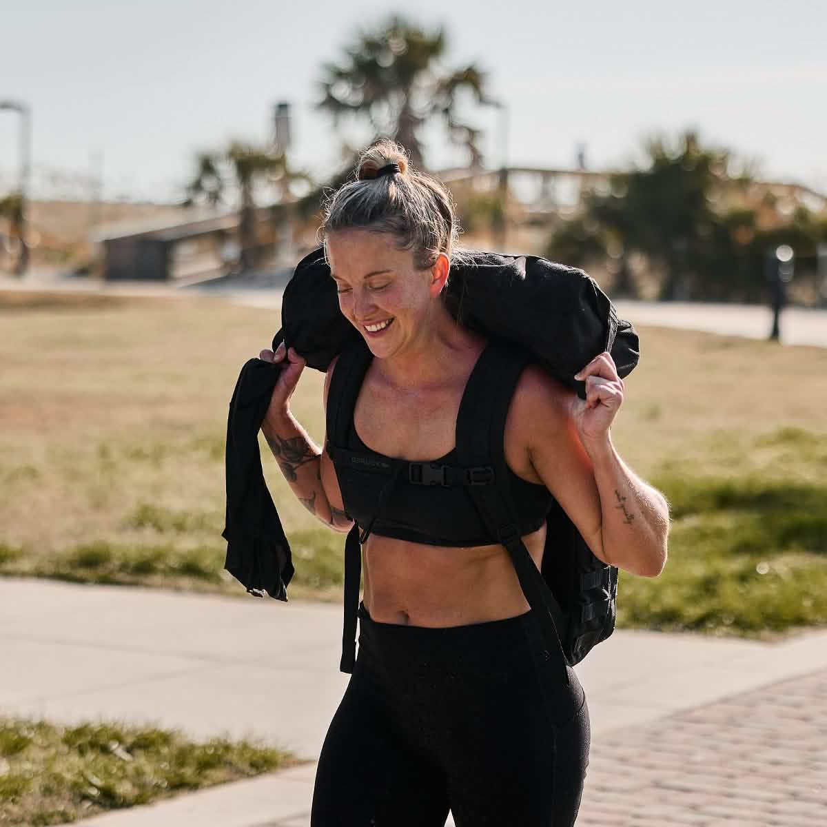 Woman rucking outdoors with GORUCK backpack, wearing athletic gear and smiling