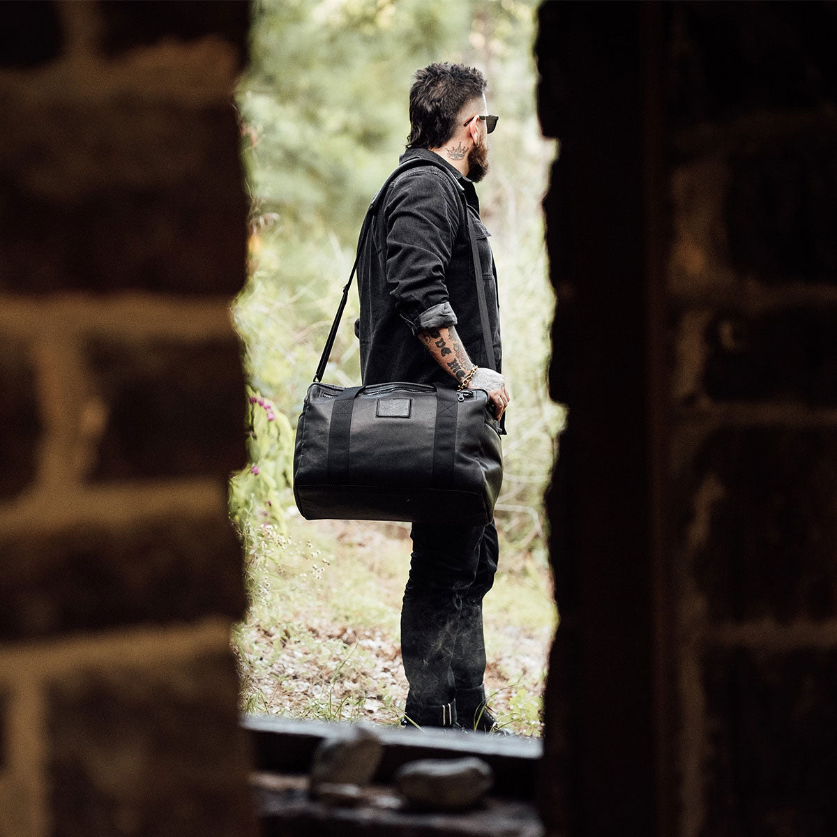 A man in dark clothes and sunglasses stands outdoors, holding a black duffel bag over his shoulder.