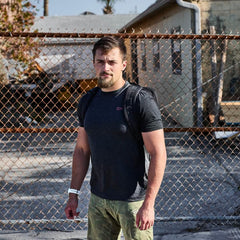 Man wearing GORUCK backpack and dark tee stands outdoors by chain-link fence