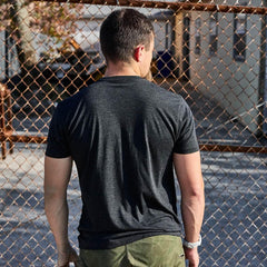 Man wearing GORUCK black athletic tee and camo shorts stands by chain link fence outdoors